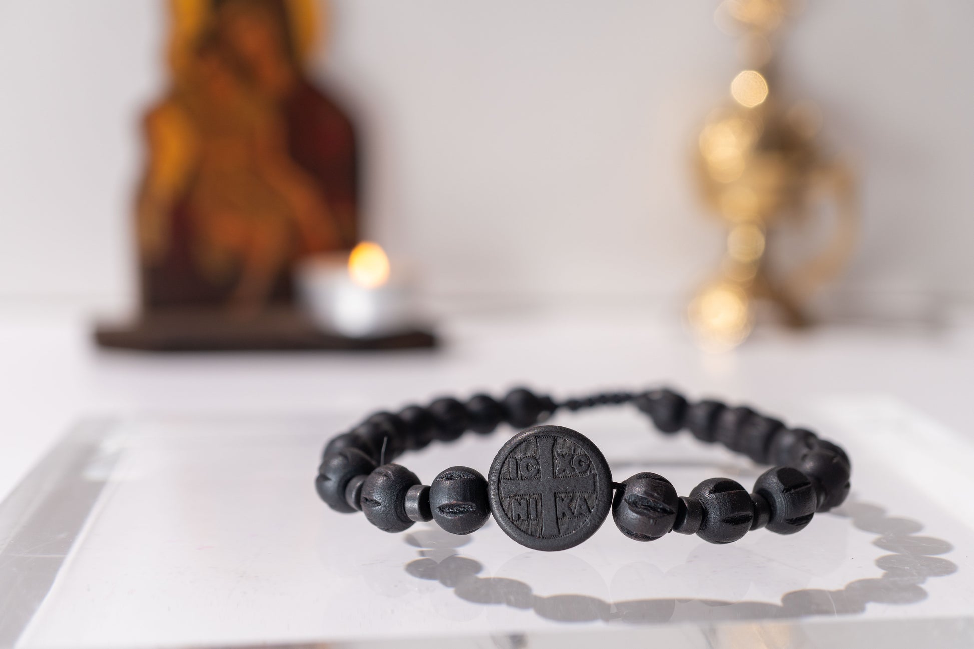 Black beaded bracelet on a white surface with blurred religious statue and candle in the background