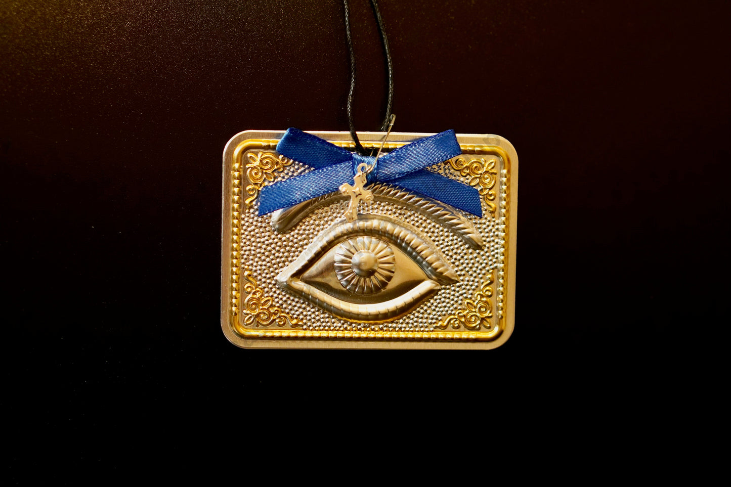 Tama votive offering featuring an embossed silver eye with gold-plated borders, a blue bow, a miniature cross charm, a hanging cord, and a safety pin.