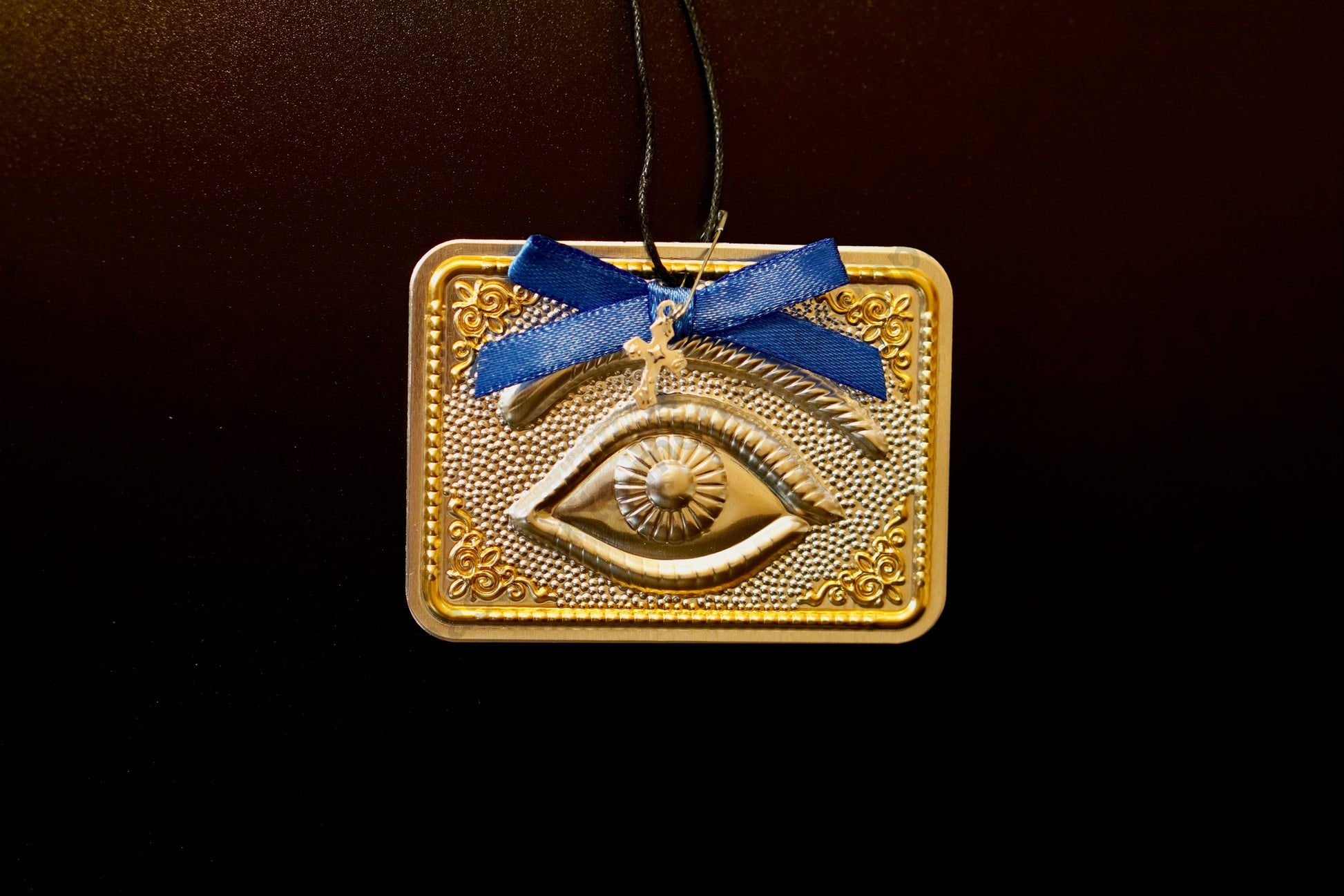 Tama votive offering featuring an embossed silver eye with gold-plated borders, a blue bow, a miniature cross charm, a hanging cord, and a safety pin.