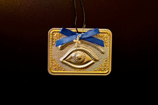 Tama votive offering featuring an embossed silver eye with gold-plated borders, a blue bow, a miniature cross charm, a hanging cord, and a safety pin.