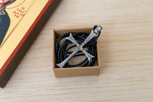 A silver-toned cross pendant necklace on a black cord, presented in a small brown cardboard box on a light wood surface.