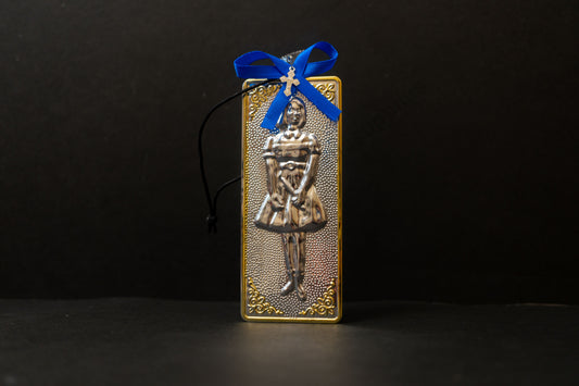 Tama votive offering featuring an embossed figure of a silver girl with gold-plated borders, a blue bow, a miniature cross charm, a hanging cord, and a safety pin.