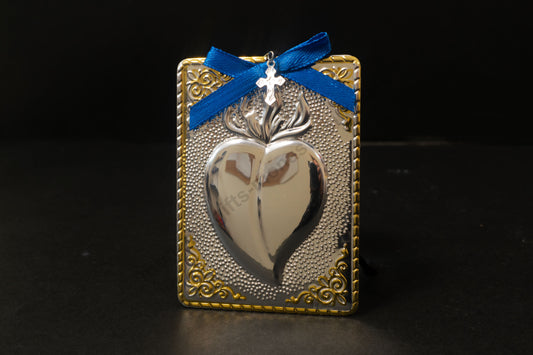 Tama votive offering featuring an embossed silver heart with gold-plated borders, a blue bow, a miniature cross charm, a hanging cord, and a safety pin.