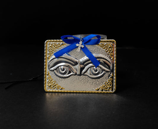 Tama votive offering featuring an embossed pair of silver eyes with gold-plated borders, a blue bow, a miniature cross charm, a hanging cord, and a safety pin.