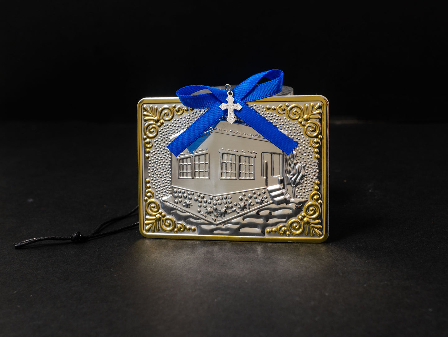 Tama votive offering featuring an embossed silver house with gold-plated borders, a blue bow, a miniature cross charm, a hanging cord, and a safety pin.
