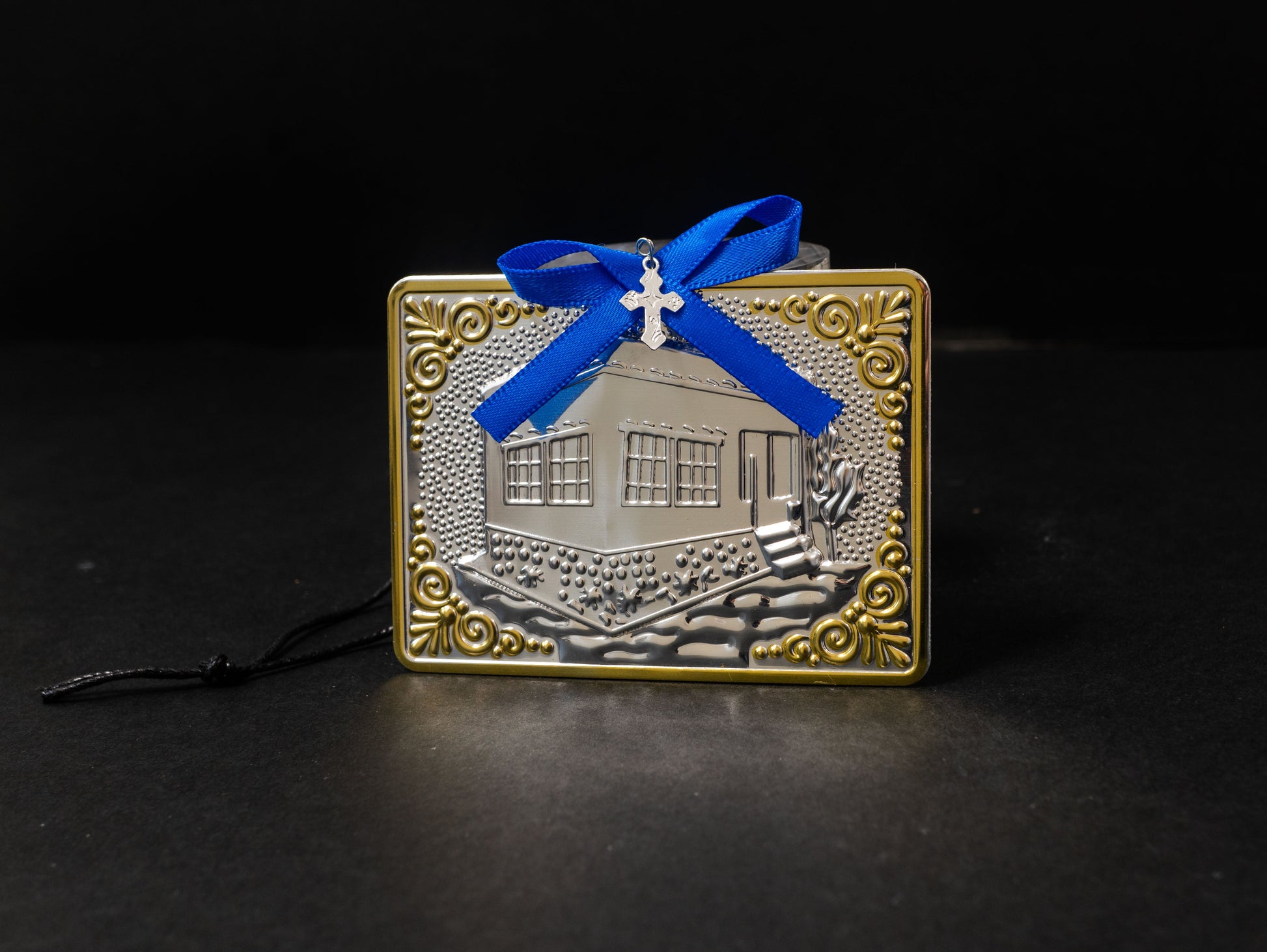 Tama votive offering featuring an embossed silver house with gold-plated borders, a blue bow, a miniature cross charm, a hanging cord, and a safety pin.