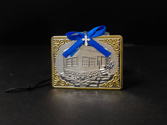 Tama votive offering featuring an embossed silver house with gold-plated borders, a blue bow, a miniature cross charm, a hanging cord, and a safety pin.