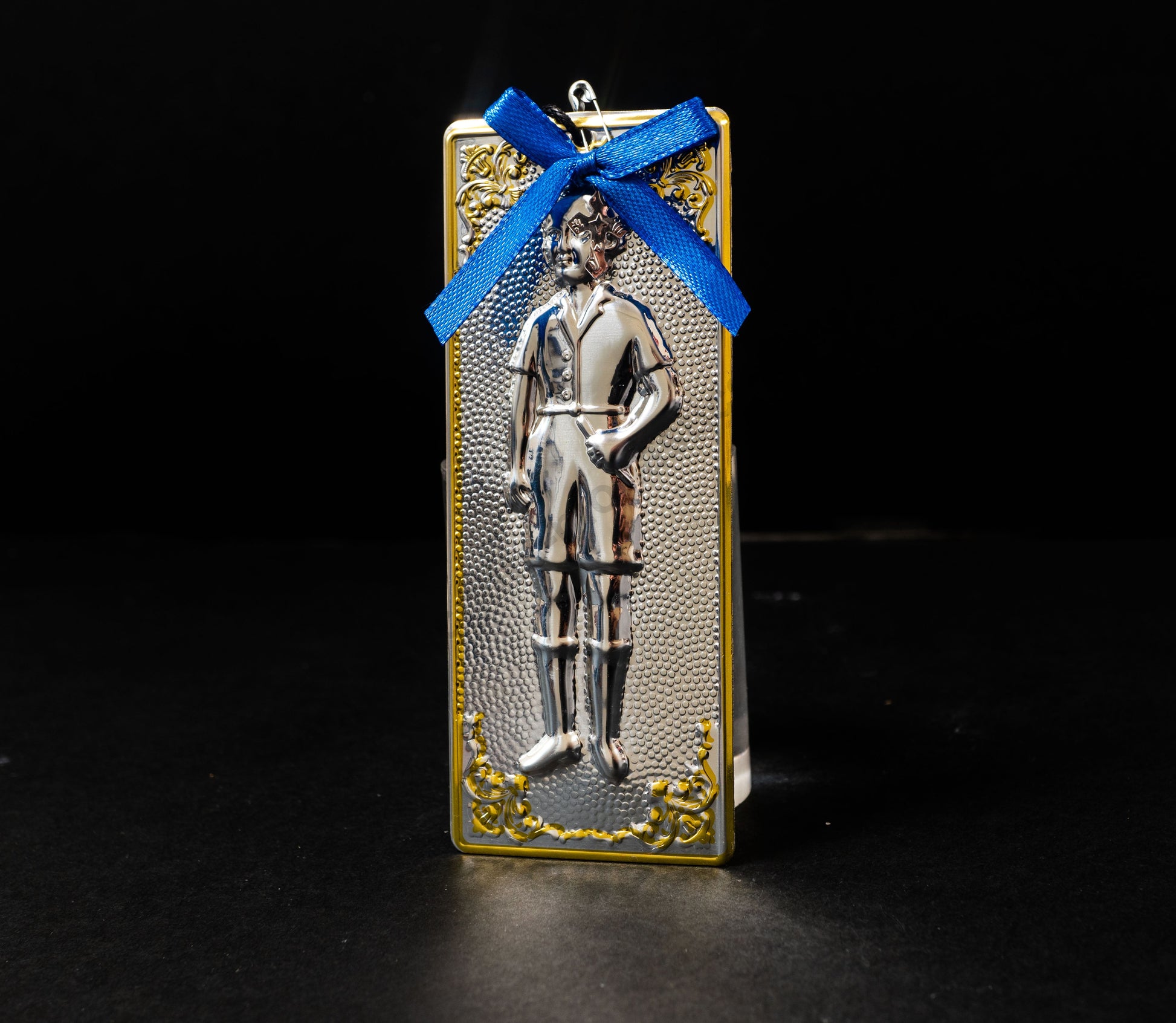 Tama votive offering featuring an embossed figure of a silver boy with gold-plated borders, a blue bow, a miniature cross charm, a hanging cord, and a safety pin.
