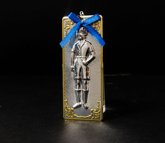 Tama votive offering featuring an embossed figure of a silver boy with gold-plated borders, a blue bow, a miniature cross charm, a hanging cord, and a safety pin.