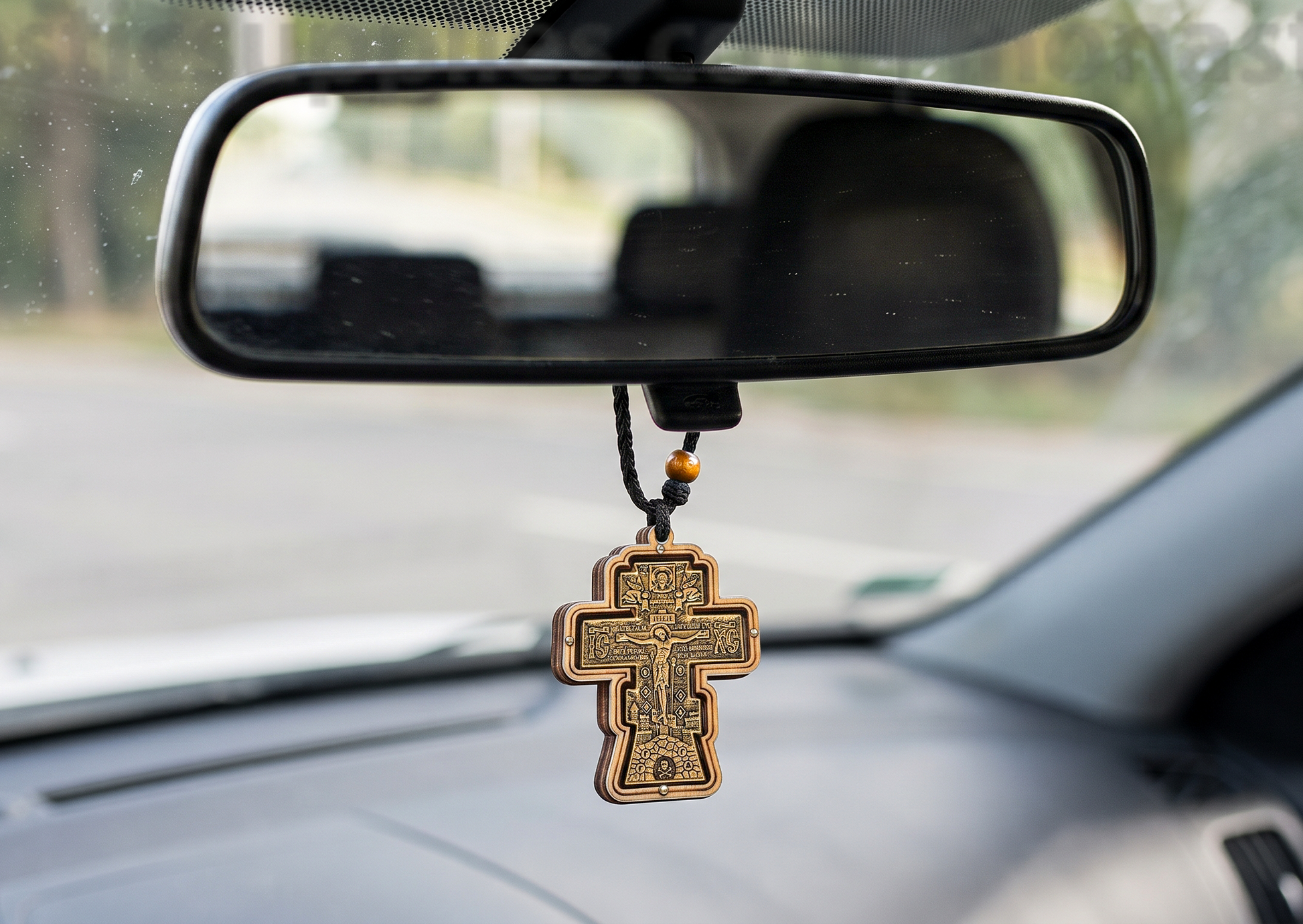 A bronze-colored Orthodox cross with a printed image of Jesus Christ hangs from a black cord beneath a car's rearview mirror.
