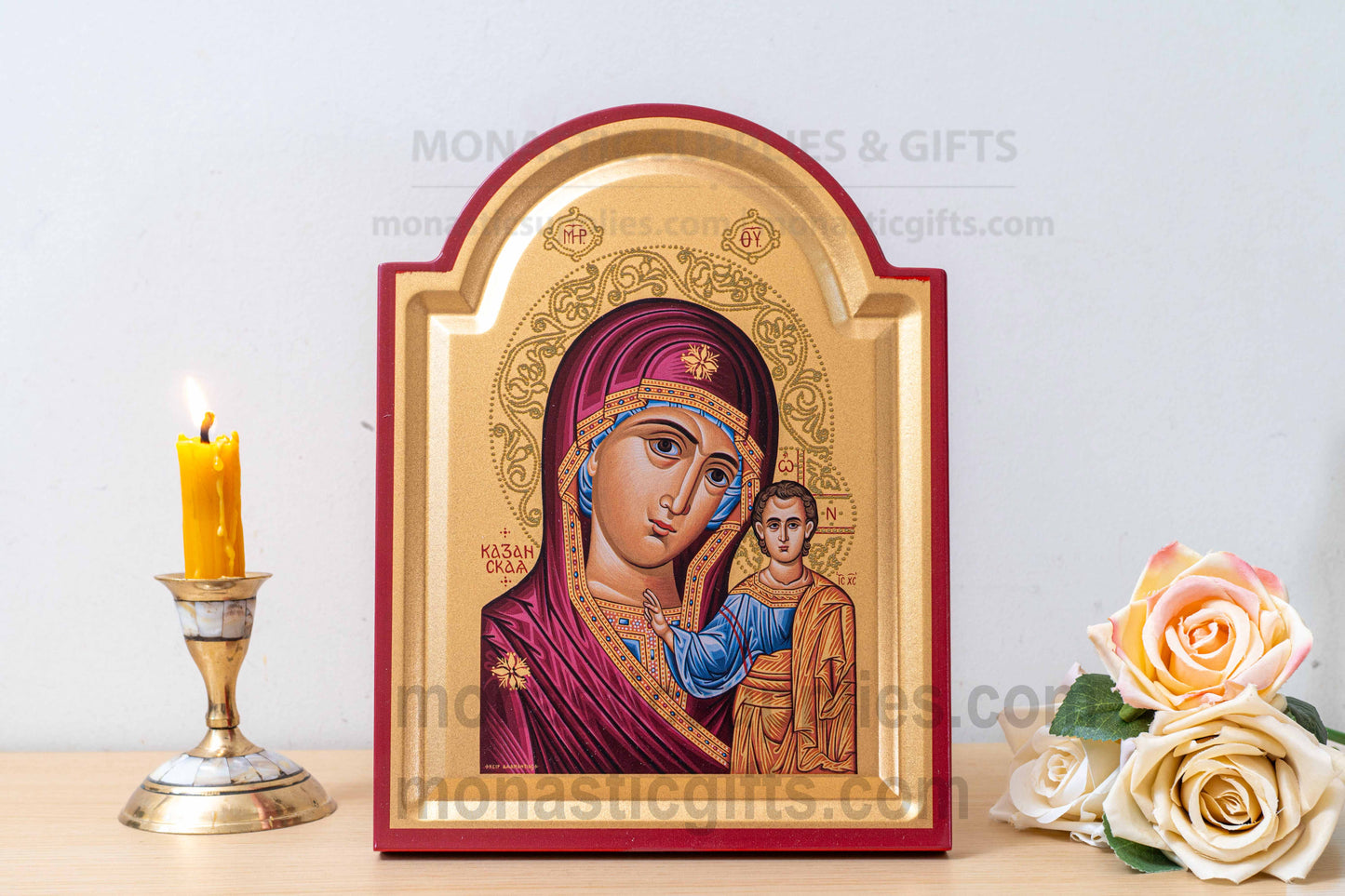 A framed Orthodox Christian icon of the Virgin Mary and Baby Jesus on a tabletop, flanked by a lit yellow candle in a brass holder on the left and artificial cream roses on the right. The icon has a gold background with a red frame.