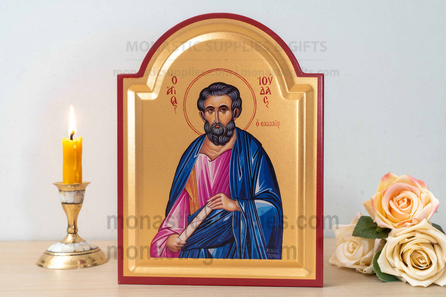 A framed religious icon of Saint Paul standing on a tabletop, flanked by a lit yellow candle in a brass holder on the left and a bouquet of pale peach roses on the right. The icon has a gold background with a red border and depicts Saint Paul in blue and pink robes.