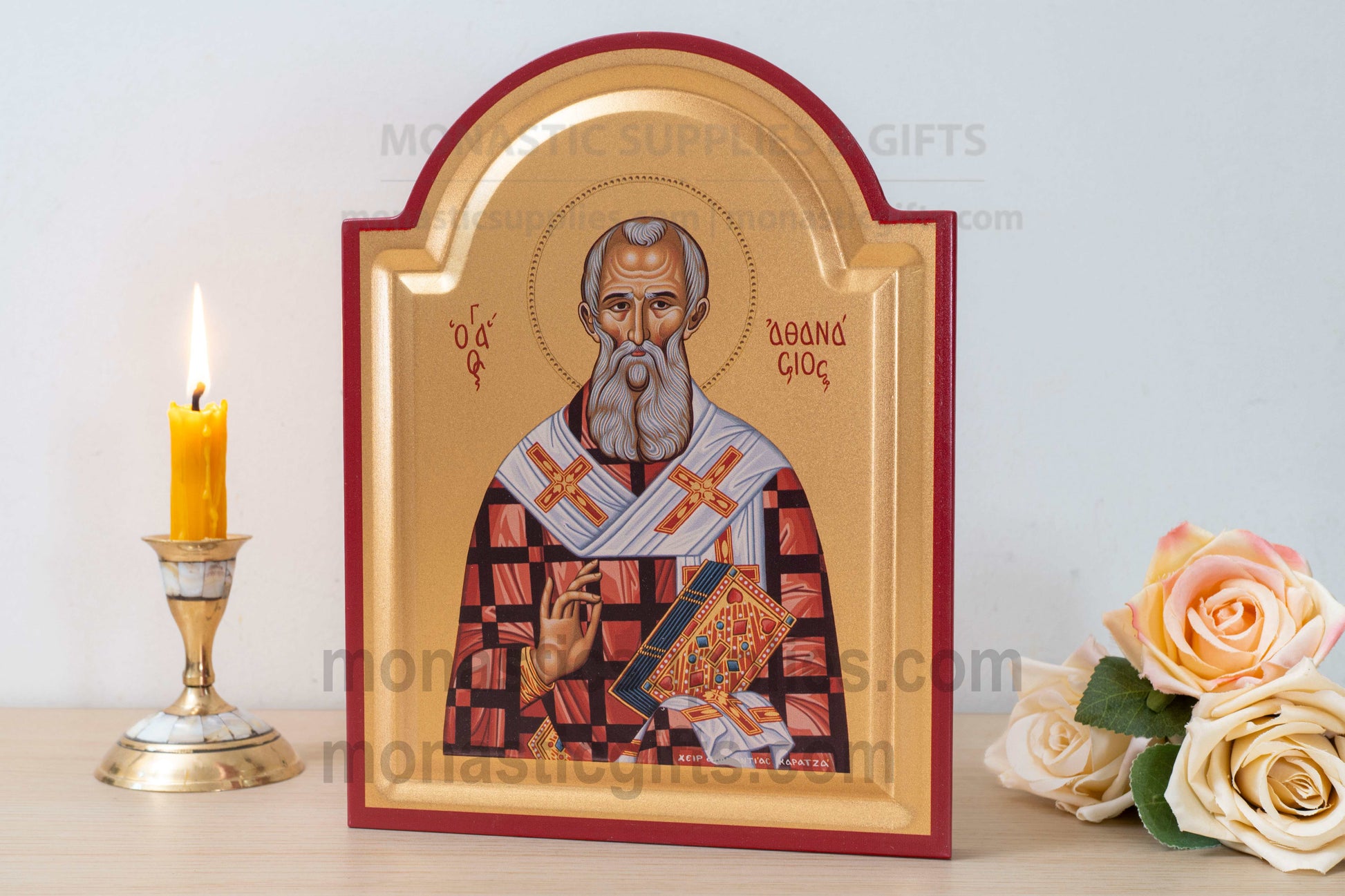 A framed religious icon of Saint George on a tabletop, styled with a lit yellow candle in a brass holder on the left and cream roses on the right. The icon has a gold background with a red arched border and depicts the saint in red, white, and blue vestments.