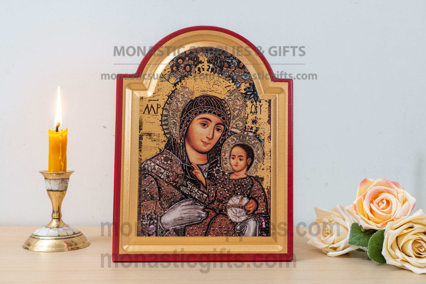 A framed Orthodox-style religious icon showing the Virgin Mary holding the Christ child, displayed on a tabletop beside a lit yellow candle in a brass holder and pale peach roses. The icon has a red border and gold-toned background, with visible watermark text across the image.