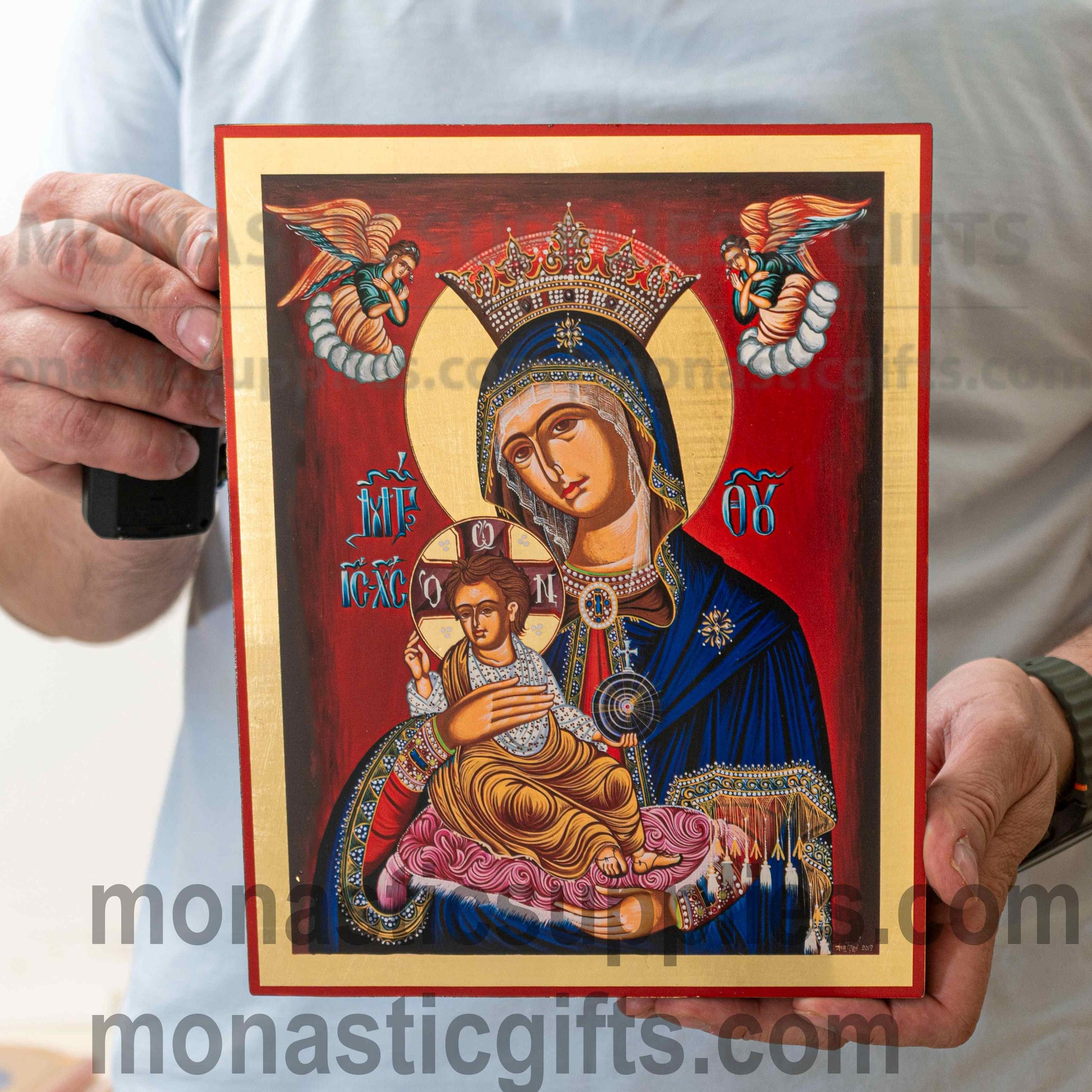 A person holds a rectangular religious icon painting featuring the Virgin Mary holding the infant Jesus, with a red background and gold border. The person is wearing a light gray T-shirt, and the icon has visible watermark text across it.