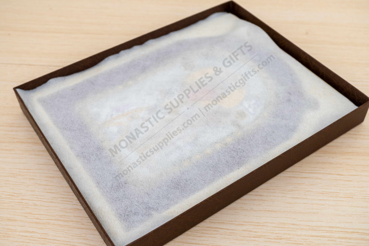 A square brown gift box containing a white tissue or paper insert printed with the Monastic Supplies & Gifts website, placed on a light wood surface.