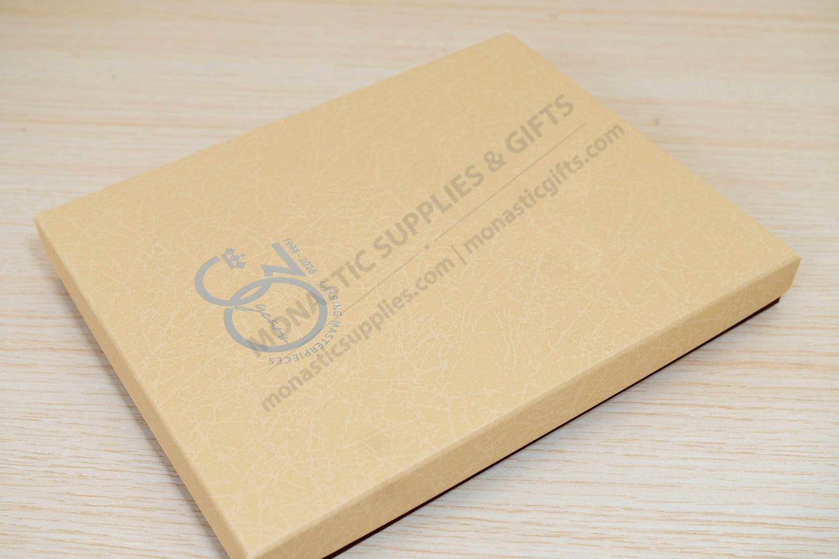 A rectangular tan gift box with a subtle embossed texture, featuring the Monastic Supplies & Gifts logo and website printed on the top, resting on a light wood grain surface.
