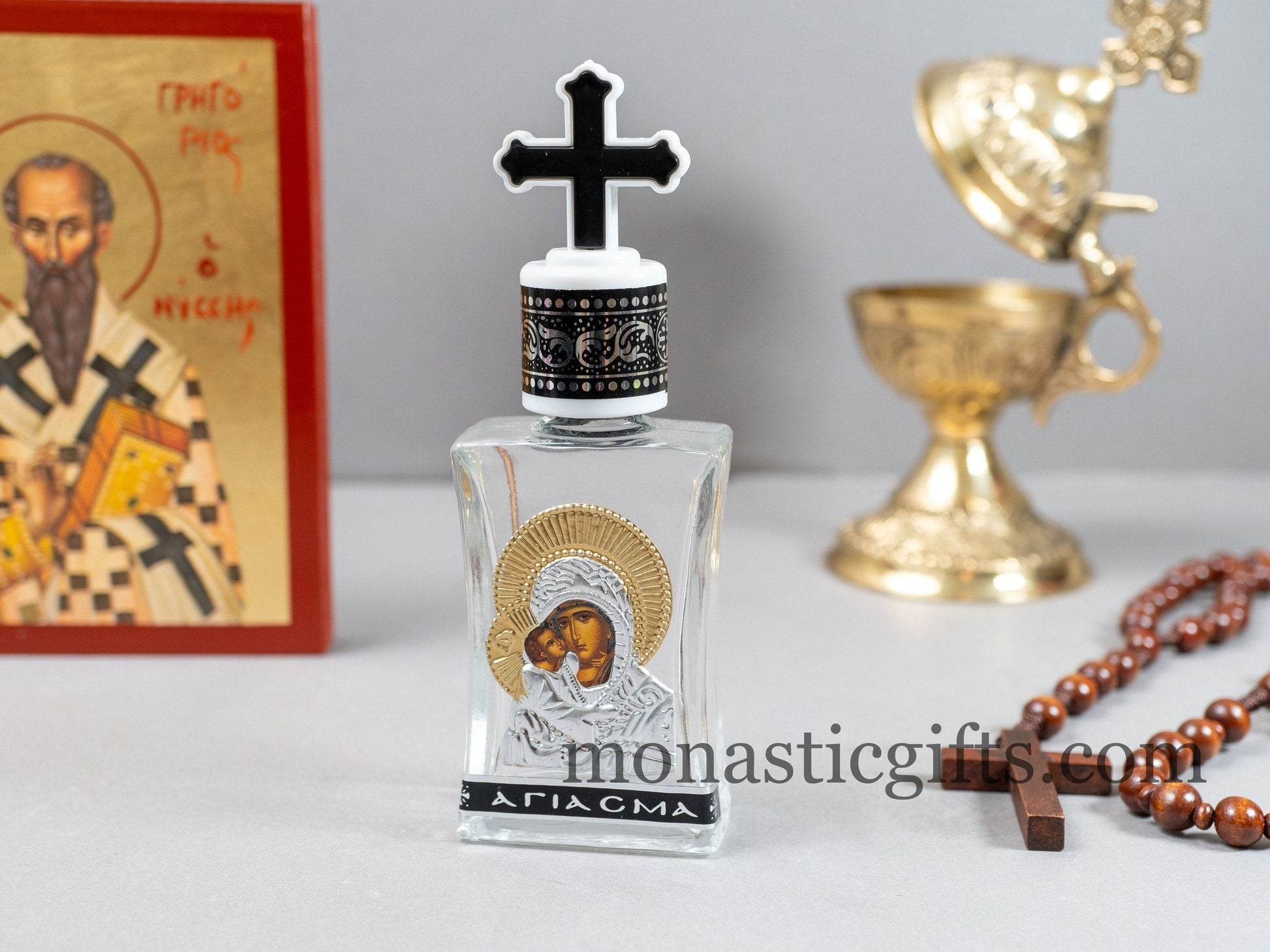 Empty clear glass Holy Water bottle featuring a silver-tone metallic icon plaque of the Panagia Theotokos and a cross-shaped screw cap.